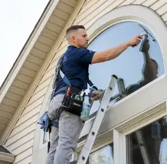 Professional window cleaner on a ladder using a squeegee to clean an exterior arched window, Albany, NY.